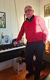 Eldre mann i rød genser står ved et elektronisk piano i en lys stue