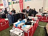 Innendørs messehall med Sport 1-stand, klær på stativer og esker på bord.
