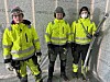 Tre personer i gule arbeidsklær står inne i et uisolert rom med blåseisolering.