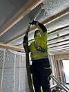 Person i verneutstyr blåseisolerer et tak i et uferdig bygg fra en stige.