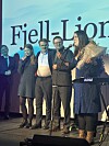 Personer på scene foran Fjell-Lion-lerret, kvinne med blomster taler ved mikrofon.