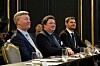 Tre menn sitter ved et bord med glass og mikrofoner i et konferanselokale.
