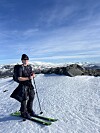 Person står på ski på en snødekt fjelltopp med fjell i bakgrunnen.