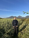En person står i høyt gress med fjell og blå himmel i bakgrunnen.