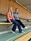 To kvinner sitter på en benk i en innendørs idrettshall med trepanel og grønt gulv.
