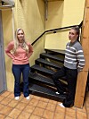 To kvinner står ved en svart trapp i en gul trappeoppgang med flislagt gulv.
